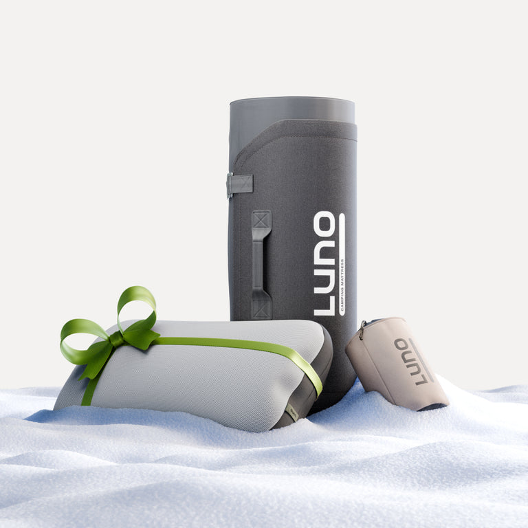 A Camping AIR+FOAM Pro Sleep Bundle by Luno® stands upright on the snowy ground, embodying car camping essentials. Beside it, a pillow wrapped with a green ribbon and a small, rolled-up beige bag create a cozy campsite feel. The background is softly muted in light gray.