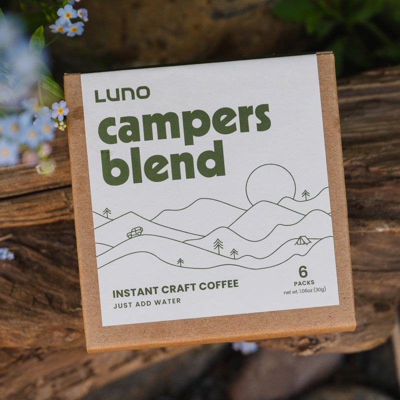 A box of Luno® Campers Blend Instant Craft Coffee is resting on a piece of wood, ready for outdoor adventures. The packaging features green text and a minimalist illustration of mountains and trees. Travel-friendly coffee at its best, the box contains 6 packs and suggests adding water to prepare.
