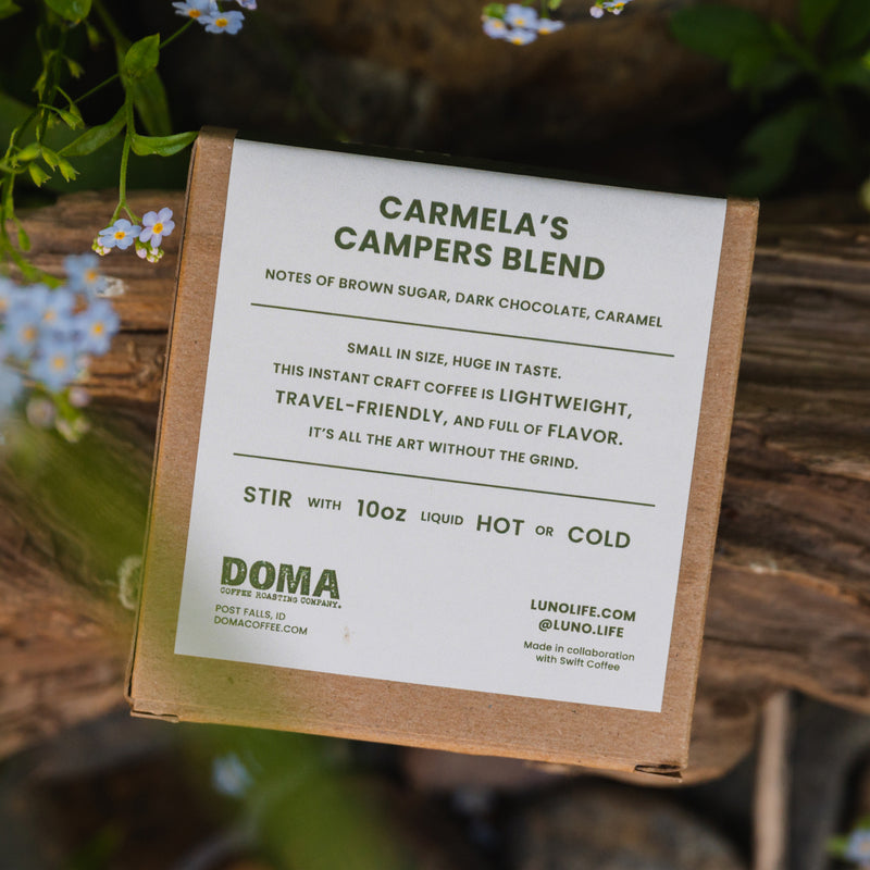 A box of Luno® Campers Blend Instant Craft Coffee titled "Campers Blend" sits on a wooden surface adorned with small white flowers. The package describes this travel-friendly coffee as having notes of brown sugar, dark chocolate, and caramel, perfect for outdoor adventures.