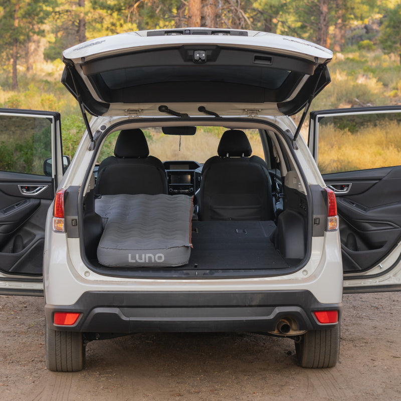 A Luno HQ AIR Vehicle Mattress (Single Side) is laid out in the cargo area of a white SUV with open doors and hatchback. Ideal for solo camping, the SUV is parked outdoors in a scenic, wooded location, providing a peaceful nature retreat.