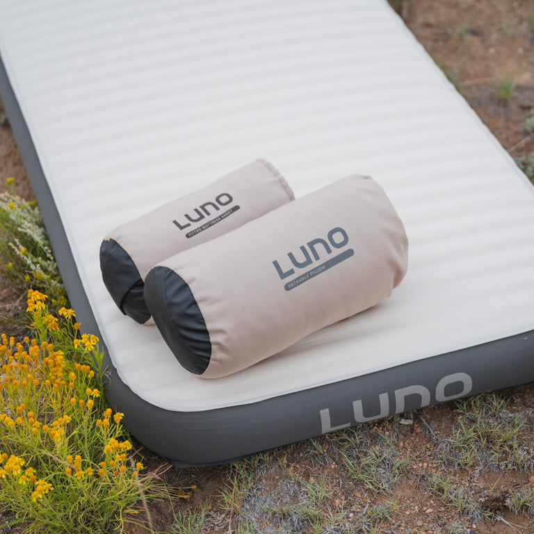 Amidst small yellow flowers and dry vegetation, the Luno® Camping AIR+FOAM Pro Sleep Bundle, which includes two rolled pillows, creates a cozy campsite vibe. The "Luno®" label denotes its status as a car camping essential, thanks to the advanced technology it incorporates.