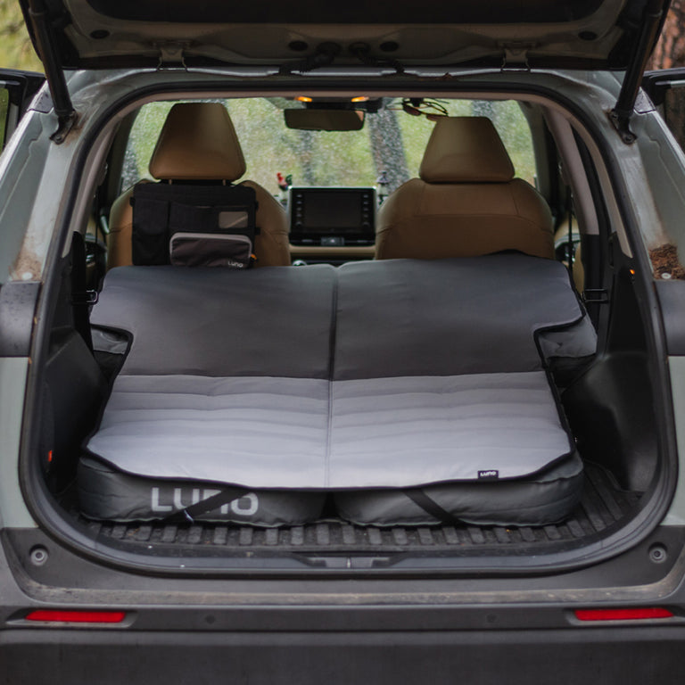 An SUV with its rear hatch open showcases the Luno® Car Camping AIR Comfy Bundle fully set up inside, covering the cargo area. The vehicle is positioned outdoors in a wooded area, visible through the windows, making it ideal for spontaneous adventures into nature.