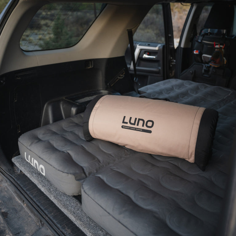 A Car Camping AIR Comfy Bundle by Luno® is set up in the back of a vehicle, with a rolled-up carry bag resting on top. The interior showcases a cozy travel arrangement, hinting at adventure-ready nights.