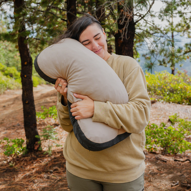 At a forest campsite, a person hugs a Luno® Camper Van AIR Starter Bundle pillow—one of their van sleeping essentials—smiling contentedly with eyes closed, dressed in a tan sweatshirt and green pants.