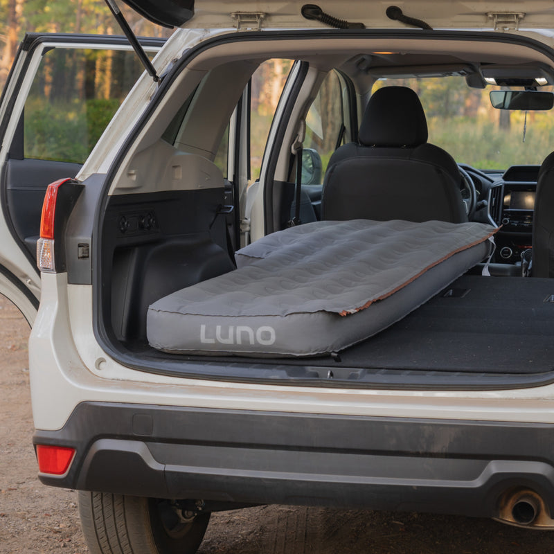 An SUV with an open hatchback features a Luno HQ AIR Vehicle Mattress (Single Side) inflated inside, ideal for solo camping trips. Parked in nature, this setup promises comfort and invites adventure.