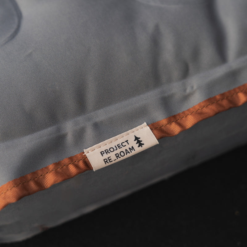 Close-up of a fabric tag on a grey AIR Vehicle Mattress (Single Side) by Luno HQ, reading "PROJECT RE-ROAM" beside a small tree icon. The mattress, upcycled with care, features an orange seam detail.
