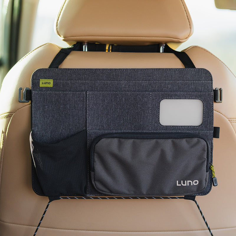 The pre-owned Seatback Organizer, branded "Luno HQ," attaches to a tan car seat, featuring a main compartment with a transparent window, mesh side pocket, and zippered pouch. Its dark gray design is available for convenient USA shipping.
