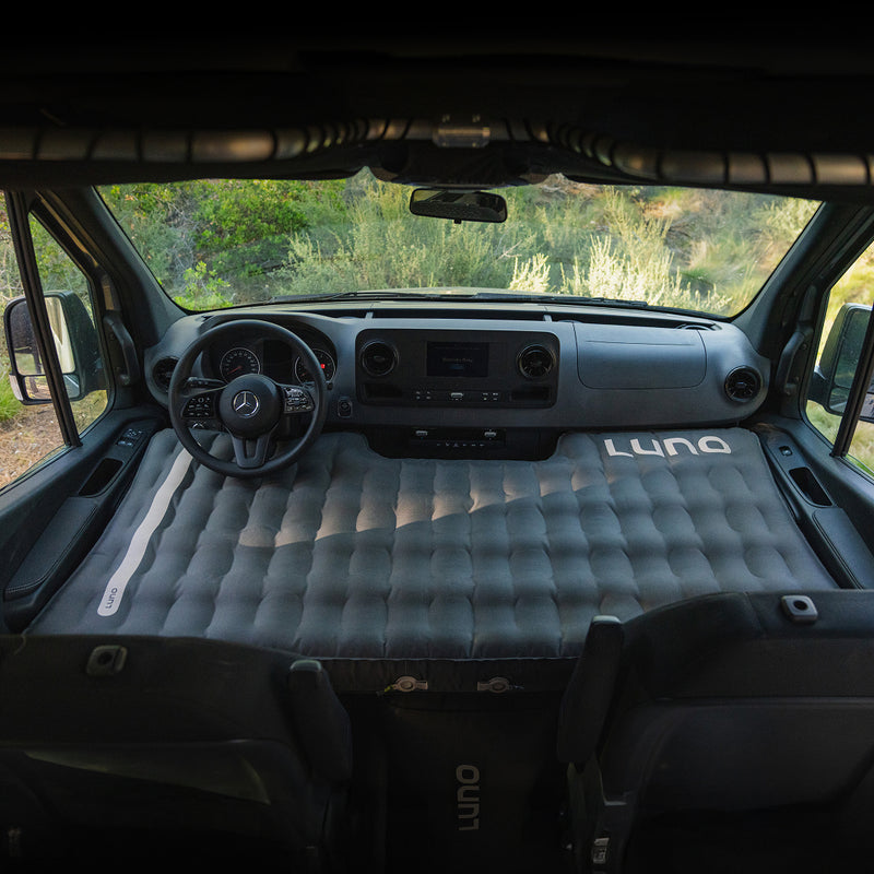 View from the back of a van featuring the Luno® Camper Van AIR Starter Bundle inflated across the front seats and dashboard, with steering wheel and windshield framing outdoor greenery.