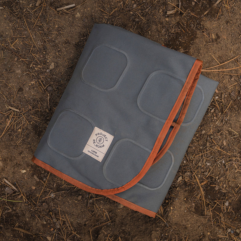 A Large Outdoor Ground Mat by Luno HQ, gray with orange trim and a white "PROUDLY USA-TOJAN" label, is folded and placed on dry brown dirt scattered with pine needles.