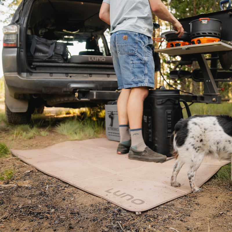 A person in shorts and socks stands by an open SUV trunk filled with camping gear, setting up a table. A small black and white dog walks on the Luno Camp Ground Mat (3x5’) in a forest outdoor setting.