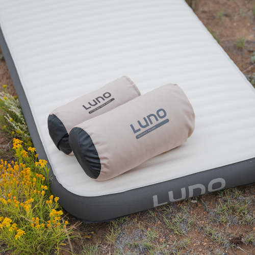 A gray inflatable mattress rests on the grassy ground, perfectly complemented by two rolled-up air pillows from the Camping AIR+FOAM Pro Sleep Bundle. The pillows, featuring their beige covers with Luno® branding, add a touch of comfort to this camping setup. Small yellow flowers peek through at the side.