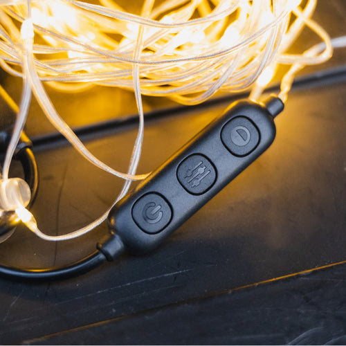 Close-up of a black corded remote with three buttons, surrounded by a tangle of warm white string lights. The buttons feature symbols for power, mode, and brightness adjustment. The cord and lights sit on a dark surface.