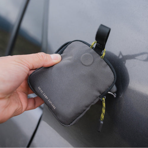 A hand is holding a small, gray, zippered pouch with a loop and printed text that reads "DRIVE CAMP EXPLORE." The pouch is attached to a car door handle using the loop. The background shows part of a car door.