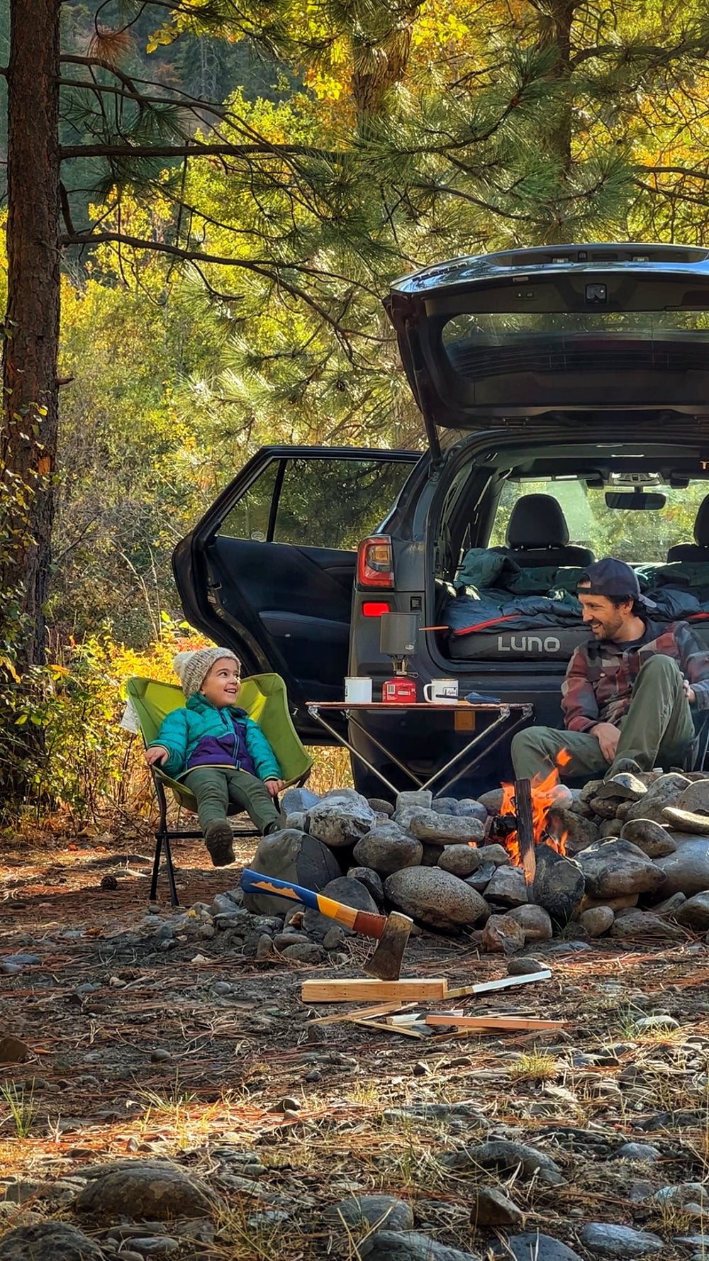 A man and a child sit on camping chairs around a small campfire. They are in a wooded area with pine trees. Behind them, an SUV with an open trunk is parked, showing camping gear inside. The scene depicts a cozy camping moment surrounded by nature.