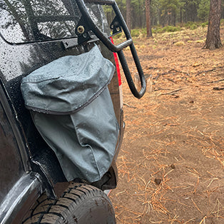 A close-up view of a black vehicle's rear exterior with a blue-gray storage bag attached, partially covered by a metal guard. The scene is set in a rainy, wooded area with tall trees and wet ground covered in pine needles.