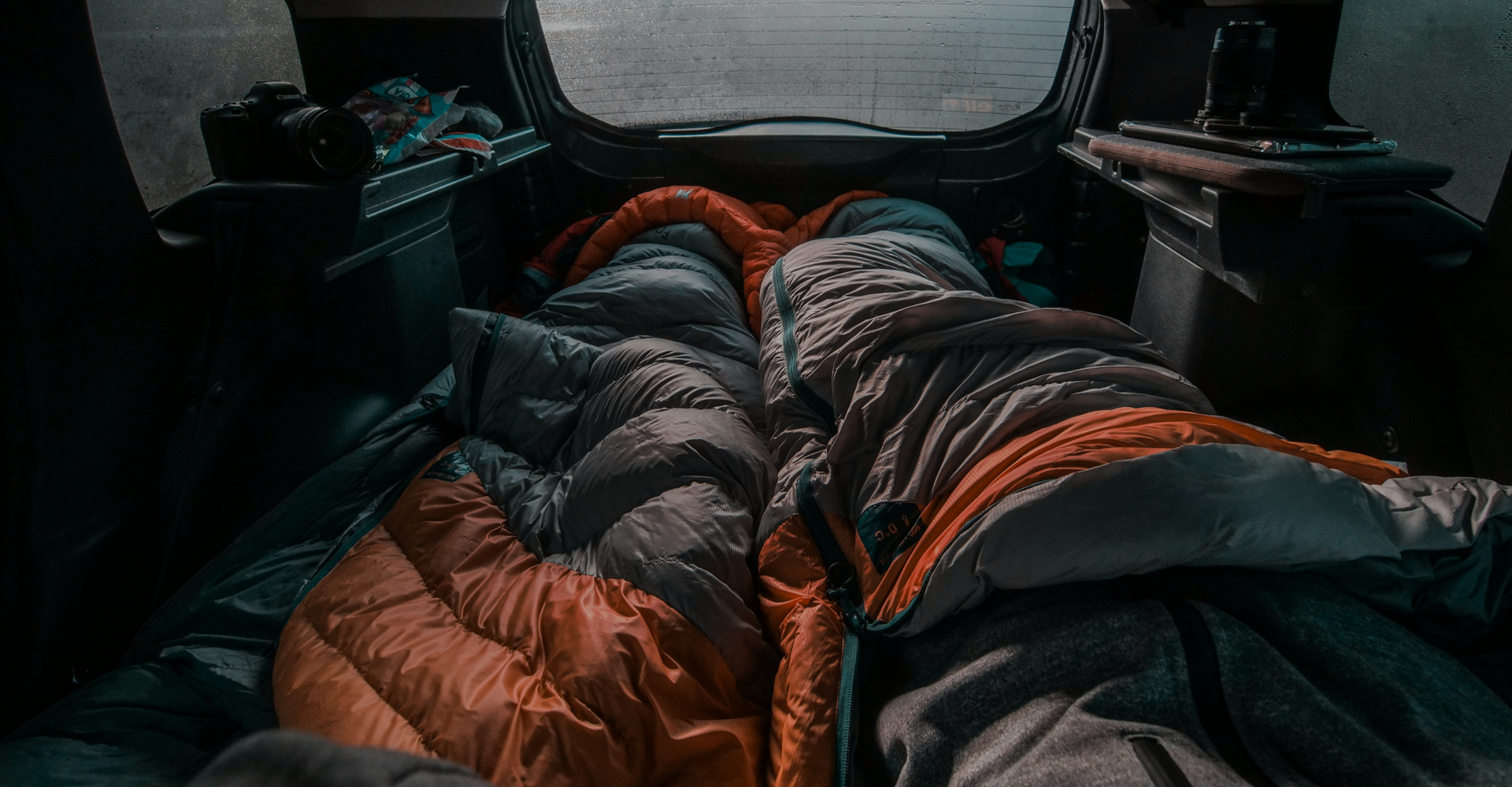 Two people are lying inside the back of a car, wrapped in orange and grey sleeping bags. The interior of the car is dimly lit, with blurred windows showing a misty outside. Camping gear and a camera are visible in the surrounding area.