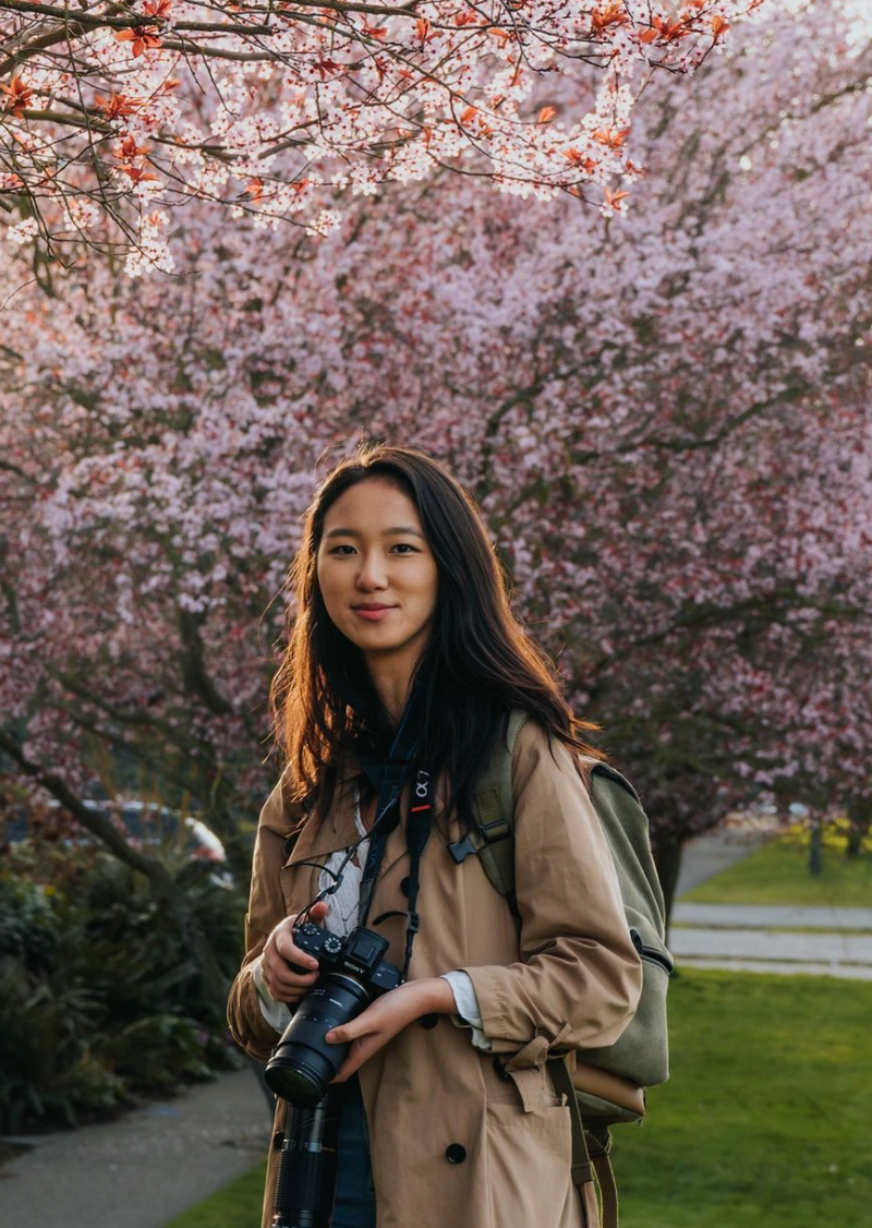 A young woman with long dark hair stands outdoors, wearing a beige coat and carrying a green backpack. She holds a professional camera and is surrounded by blossoming cherry trees, creating a vibrant and serene atmosphere.