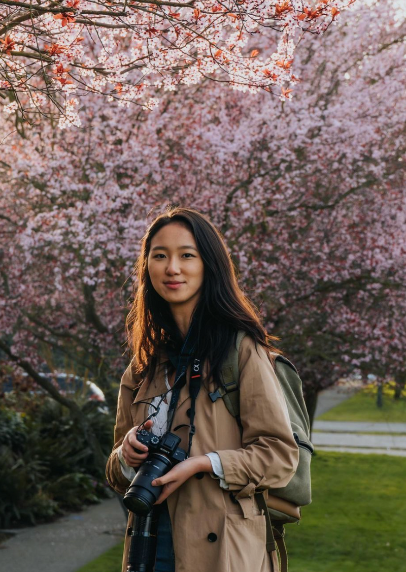 A person with long dark hair stands outdoors, holding a camera and wearing a beige trench coat and a backpack. Behind them are blooming cherry blossom trees and a park path. The scene is bathed in soft, natural light.