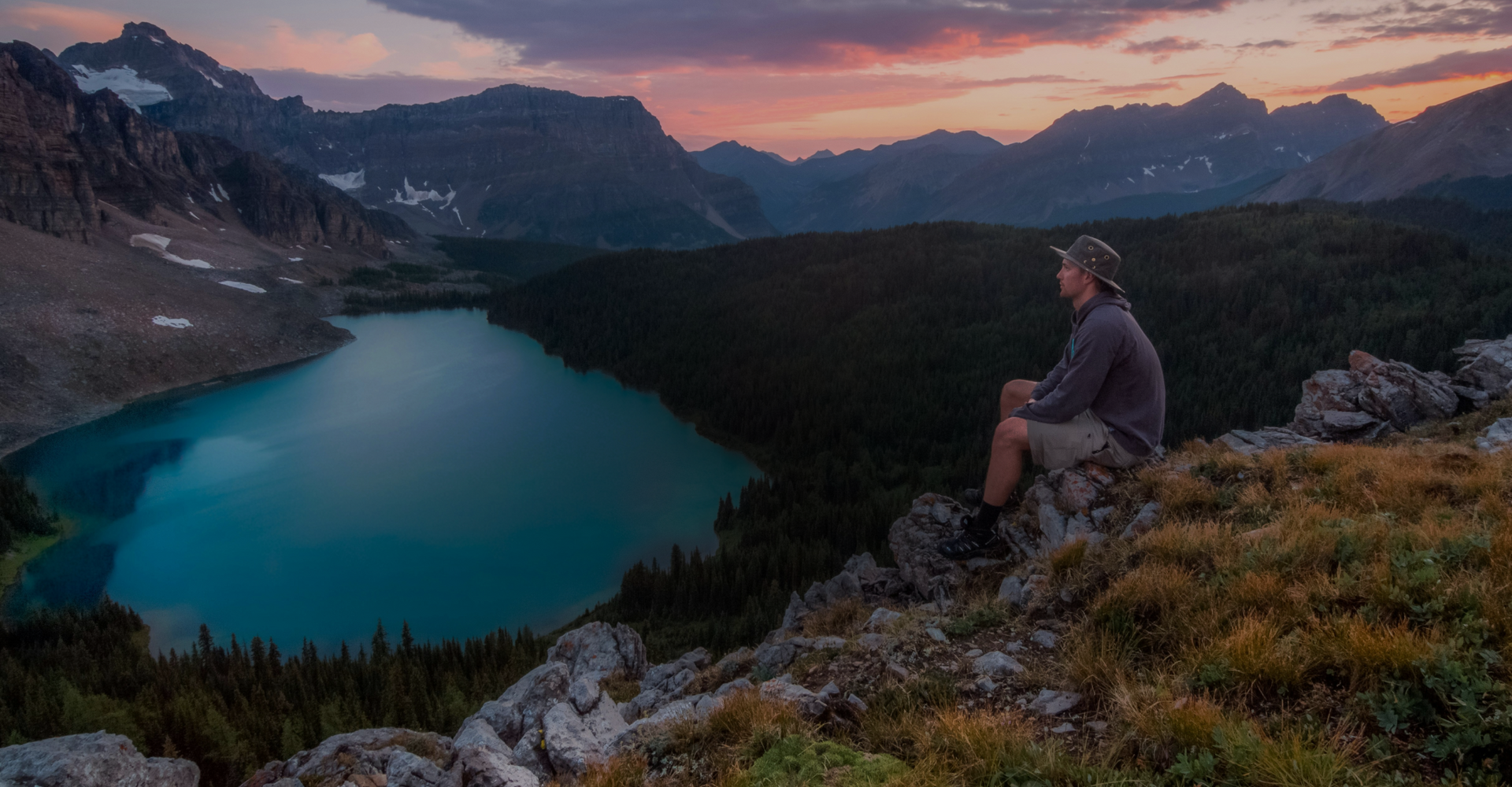 A person wearing a hat and outdoor clothing, sits on a rocky ledge overlooking a serene mountain lake surrounded by forested hills. The sky is colorful with hues of pink, orange, and purple, indicating either sunrise or sunset. Snow-capped peaks are visible in the distance.