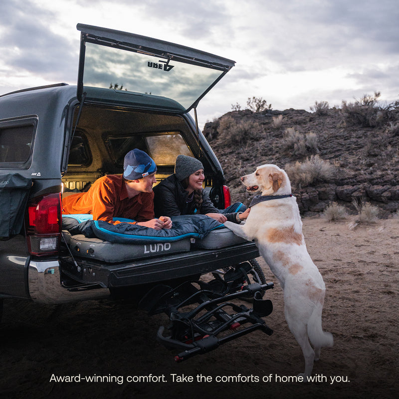 Two people relax in the bed of a truck on a Luno® AIR Truck Bed Mattress with blankets while a large dog stands on its hind legs to interact. The truck is parked outdoors near rocks and shrubs at sunset.