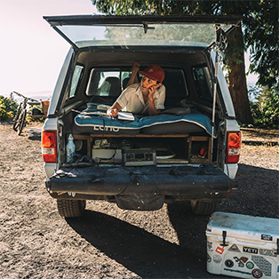 A person in a red cap lies inside the open bed of a truck, propped up on one elbow, looking contemplative with a hand on their face. The truck has a mattress and various belongings. A cooler with stickers sits on the ground by the truck. Trees and a bicycle are visible in the background.