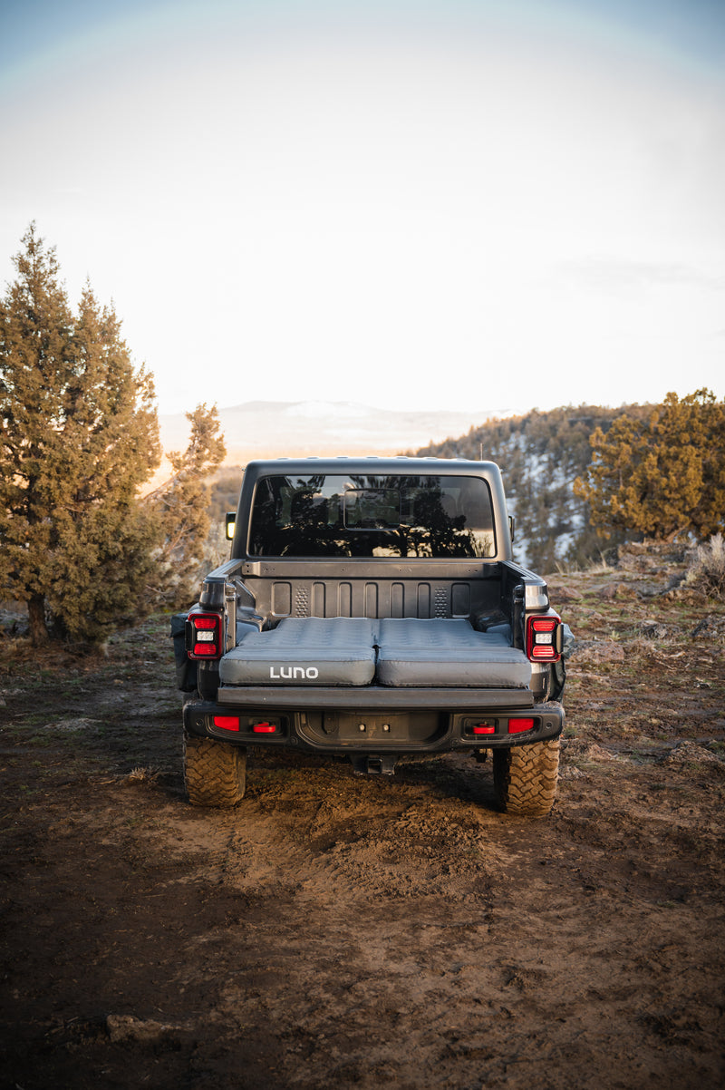 A rugged off-road vehicle parked in a scenic, mountainous area with trees around. The truck bed is open, revealing an inflatable mattress labeled "Luno," set up for camping. The sun is setting or rising, casting a warm glow on the landscape.