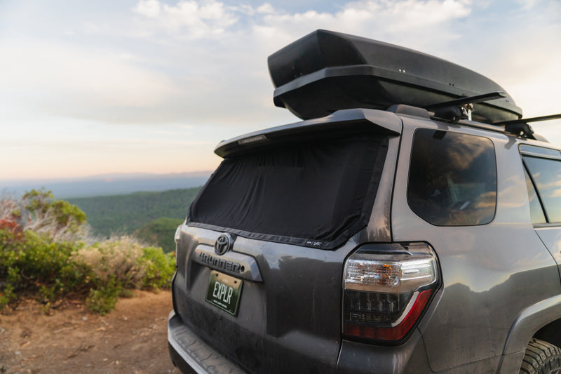 A parked SUV with a roof box and a rear window cover overlooks a scenic mountain landscape. The sky is partly cloudy, and the area is surrounded by lush greenery. The view extends into the distance, showcasing a blend of green hills and open sky.