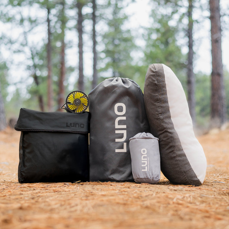 Set against a serene backdrop of tall trees and pine needles, the Camping gear showcased includes essentials for van life such as a black foldable chair, a small portable fan, and a large gray bag featuring the "Luno" logo. The Camper Van AIR Ultimate Bundle by Luno® offers added comfort with its compact gray pouch and plush pillow.