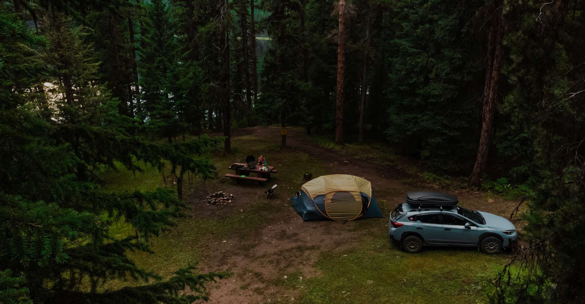 A campsite in a dense forest with a yellow and blue tent, a gray SUV nearby, and a picnic table. Two people are sitting at the table, surrounded by tall trees. There is a campfire ring on the ground and camping equipment scattered around.