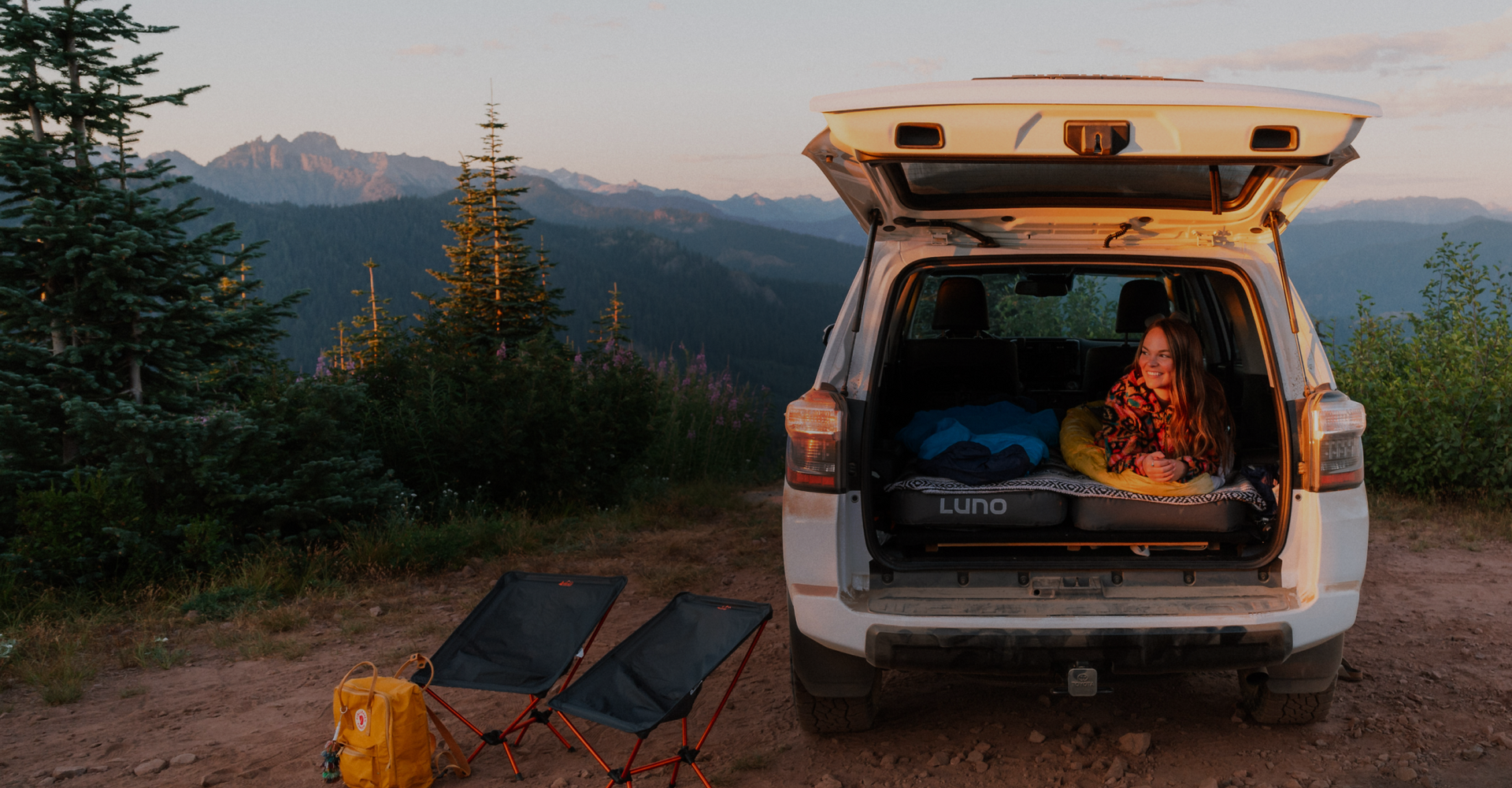 A person lounges in the back of an open SUV parked on a dirt clearing surrounded by trees and mountains. Two folding chairs and a yellow backpack are set up nearby, suggesting a camping scene. The sky is clear with a warm light, hinting at either sunrise or sunset.