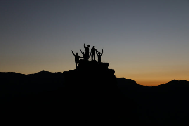 A group of people silhouetted against a sunset sky stand on a rocky peak with their hands raised in celebration. The sky has soft shades of orange and blue, and the outline of distant mountains is visible below.