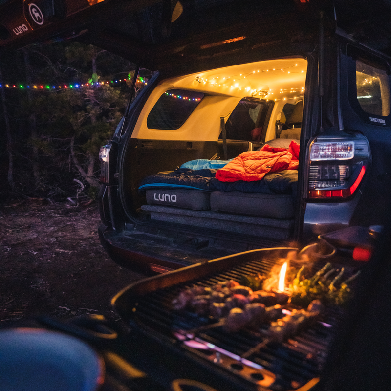 A cozy car camping setup in a dark, wooded area. The open back of an SUV reveals a sleeping area featuring the Luno® AIR Vehicle Mattress illuminated by string lights, alongside a red sleeping bag made from durable fabric. In the foreground, a grill with glowing coals is cooking food, creating a warm and inviting scene.