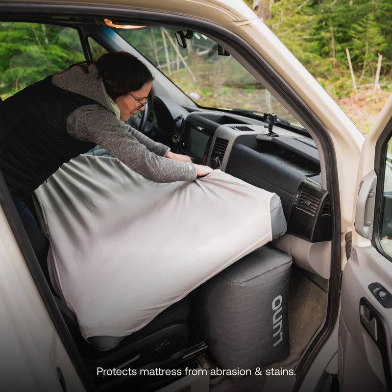 A person adjusts the Luno® AIR Van Front Cab Mattress Fitted Sheet inside a van, with the front seats visible. The van is parked in a forested area. A rolled-up temperature-regulated insulation mattress topper is on the floor. Text at the bottom reads, "Protects mattress from abrasion & stains.