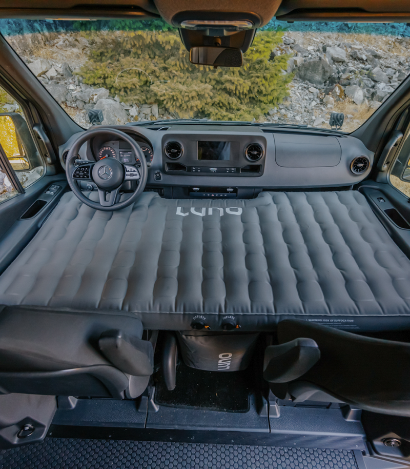 Interior view of a vehicle's front cabin with a gray inflatable mattress deployed across the driver and passenger seats, branded "Luno." The vehicle appears to be parked near a rocky and forested area, visible through the windshield.