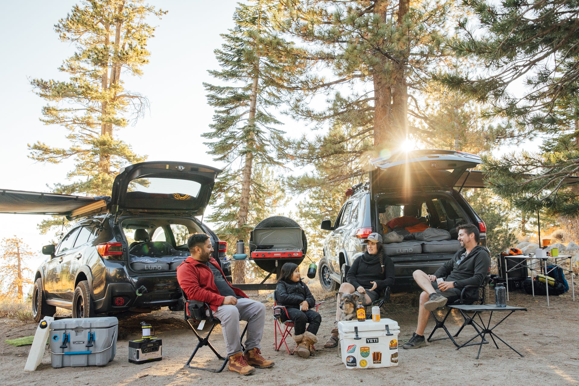 A group of four people is sitting on camping chairs around a makeshift outdoor campsite. Two SUVs with open trunks are parked nearby, and pine trees surround the area. Coolers, boxes, and camping gear are scattered around the campsite as sunlight filters through the trees.