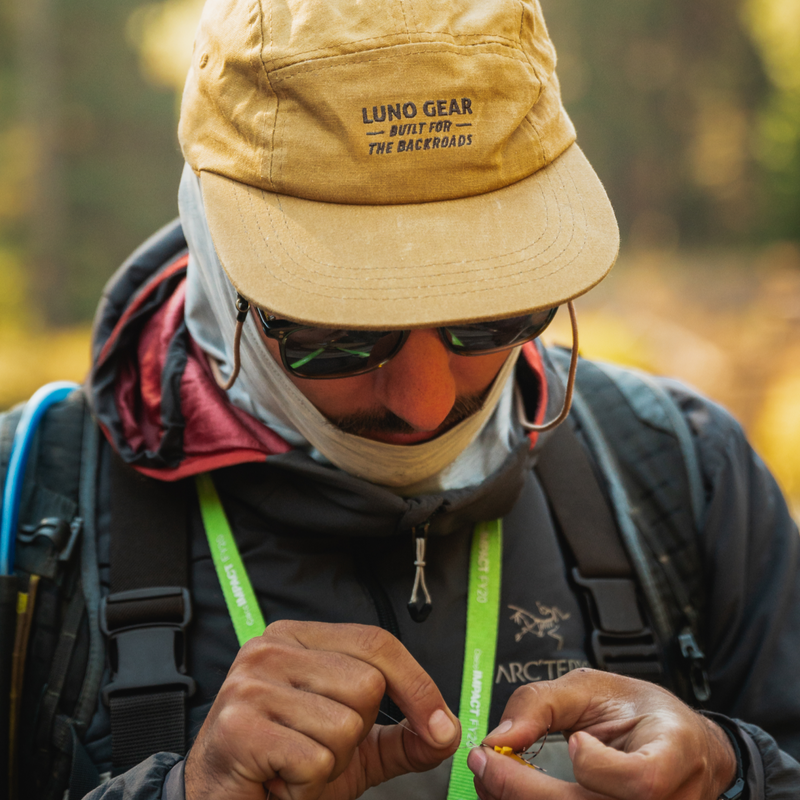 A person wearing a yellow "Backroads Canvas Hat" by Luno HQ and sunglasses is focused on an object in their hands. They have a mustache and are dressed in outdoor gear, including a grey jacket with a visible logo, and a light scarf covering their neck. The background is bright with blurred natural elements.