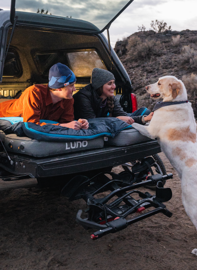 A man and woman lie in the bed of a truck, covered with a blanket and smiling at a large dog standing on its hind legs, reaching towards them. The scene takes place outdoors, with the truck's hatch open, during what appears to be a camping trip.