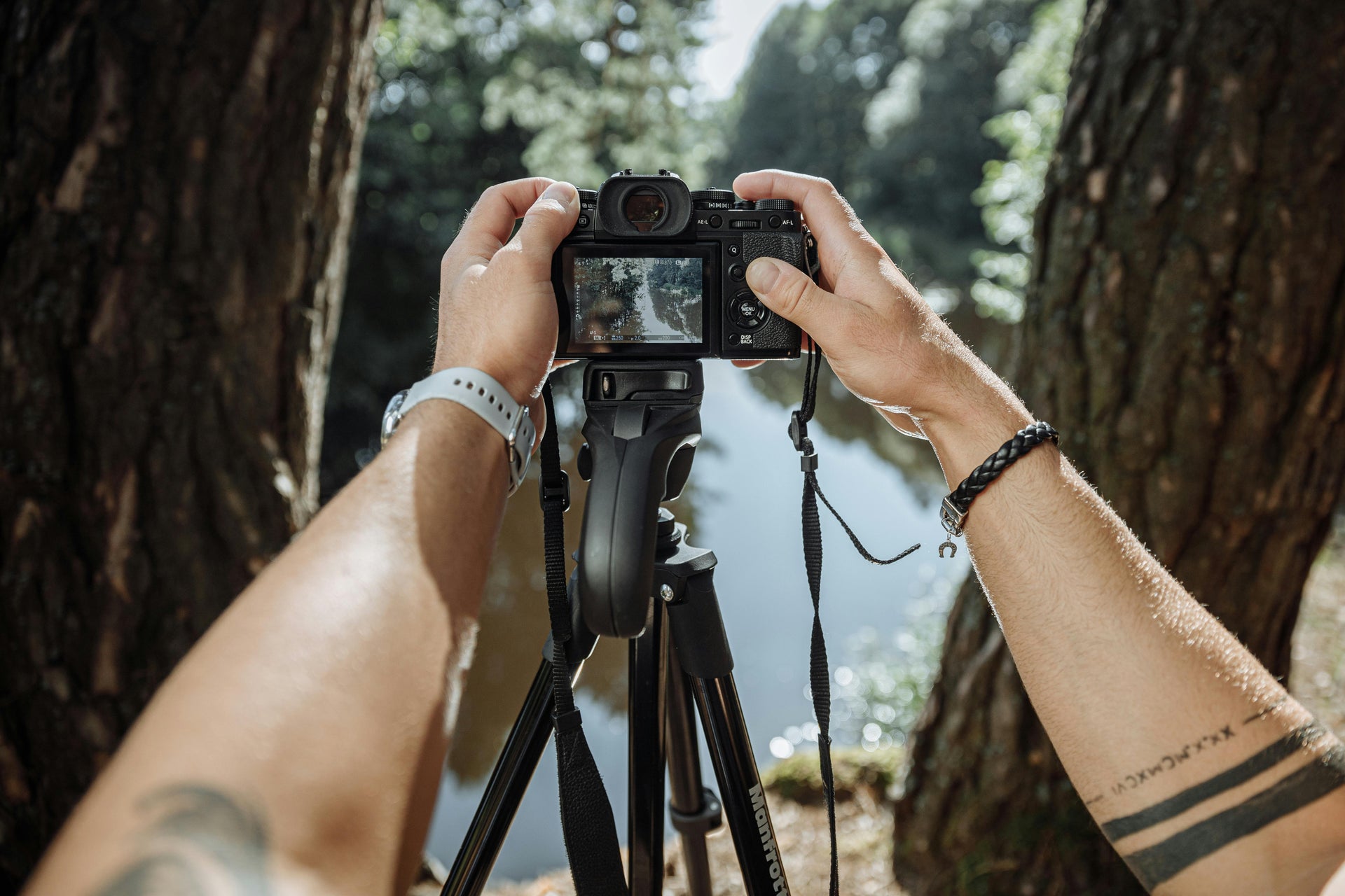 A person is taking a photo with a digital camera mounted on a tripod. The image on the camera screen shows a forested area with trees and water. The person's arms, adorned with a bracelet and a tattoo, frame the scene. Sunlight filters through the trees.