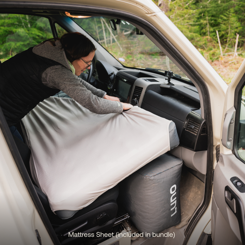 A person adjusts a mattress sheet from the "Camper Van AIR Sleep Bundle" by Luno® on the front passenger seat of a vehicle. The vehicle's interior dashboard and steering wheel are visible. The person is wearing a black vest and appears to be setting up a cozy campsite for an upcoming adventure. Text reads "Mattress Sheet (included in bundle).