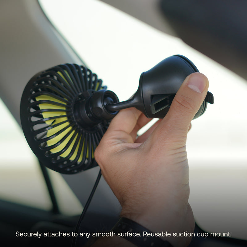 A person is securing the Luno® Car Camping Fan, showcasing its small black casing with a green interior, to a car window using the reusable suction cup mount. The fan is equipped with an articulating ball mount for optimal positioning. Caption at the bottom reads, "Securely attaches to any smooth surface. Reusable suction cup mount.