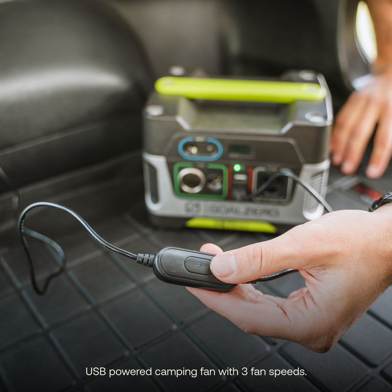 The man adjusts a small USB-powered Car Camping Fan by Luno® featuring 3 fan speeds. He is placing the fan into the USB socket on a green and gray portable power station that offers various ports and indicators. The Car Camping Fan is mounted on an articulating ball mount for flexible positioning.