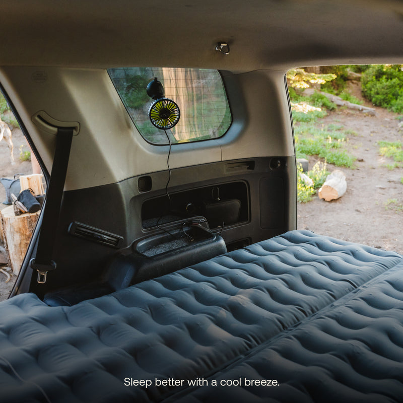 The interior of a vehicle is shown with an inflated air mattress from Luno® covering the back area. A Luno® Car Camping Fan, USB-powered, is affixed to a rear window using a stay-tight suction cup and plugged into the car's power source. The surrounding environment outside appears to be a natural setting. Text at the bottom reads, "Sleep better with a cool breeze.