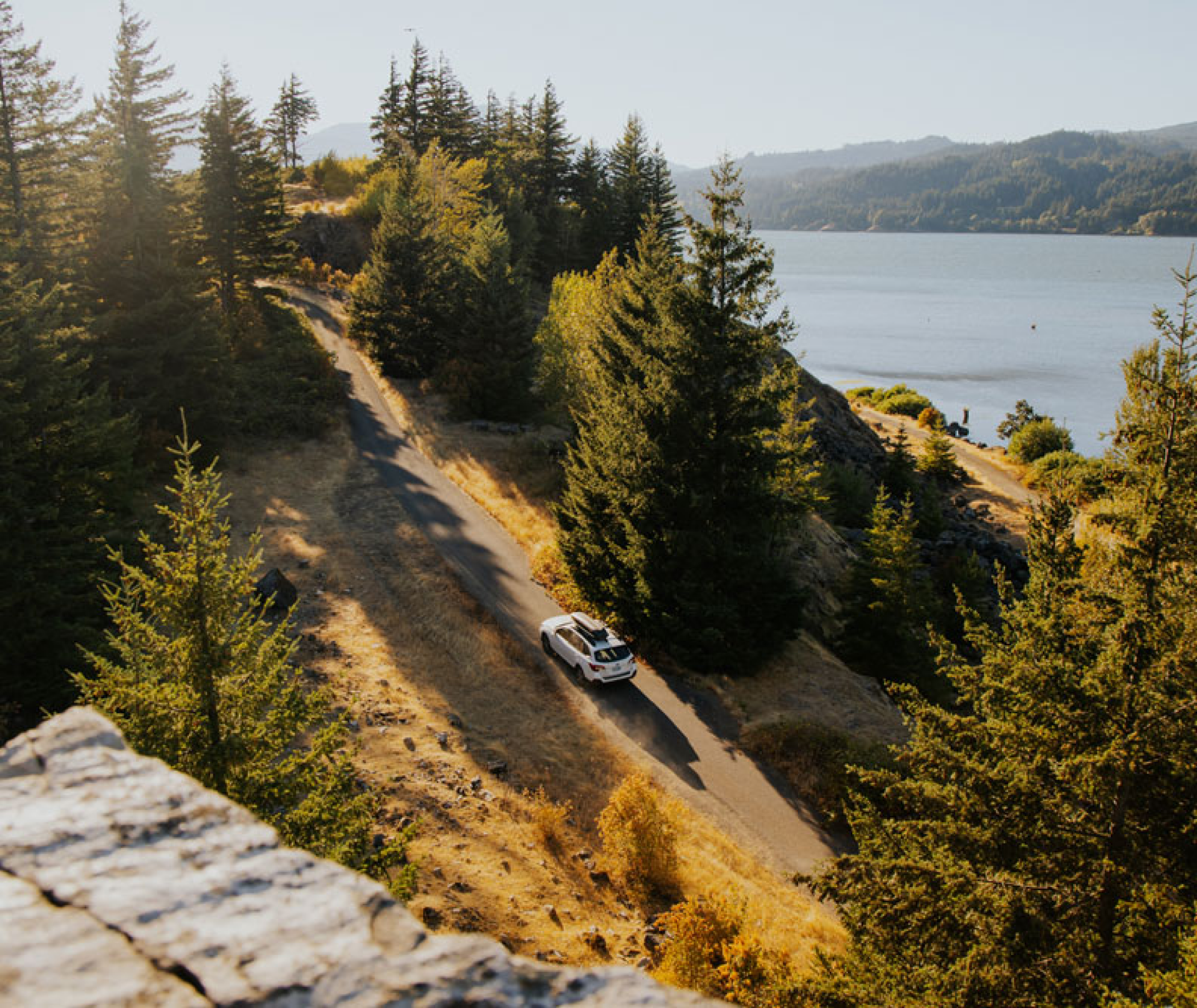 A white SUV drives along a winding road surrounded by lush green trees and dry grass. The road runs adjacent to a large body of water with mountainous terrain in the background. The scene is bathed in warm sunlight, creating a serene and picturesque landscape.