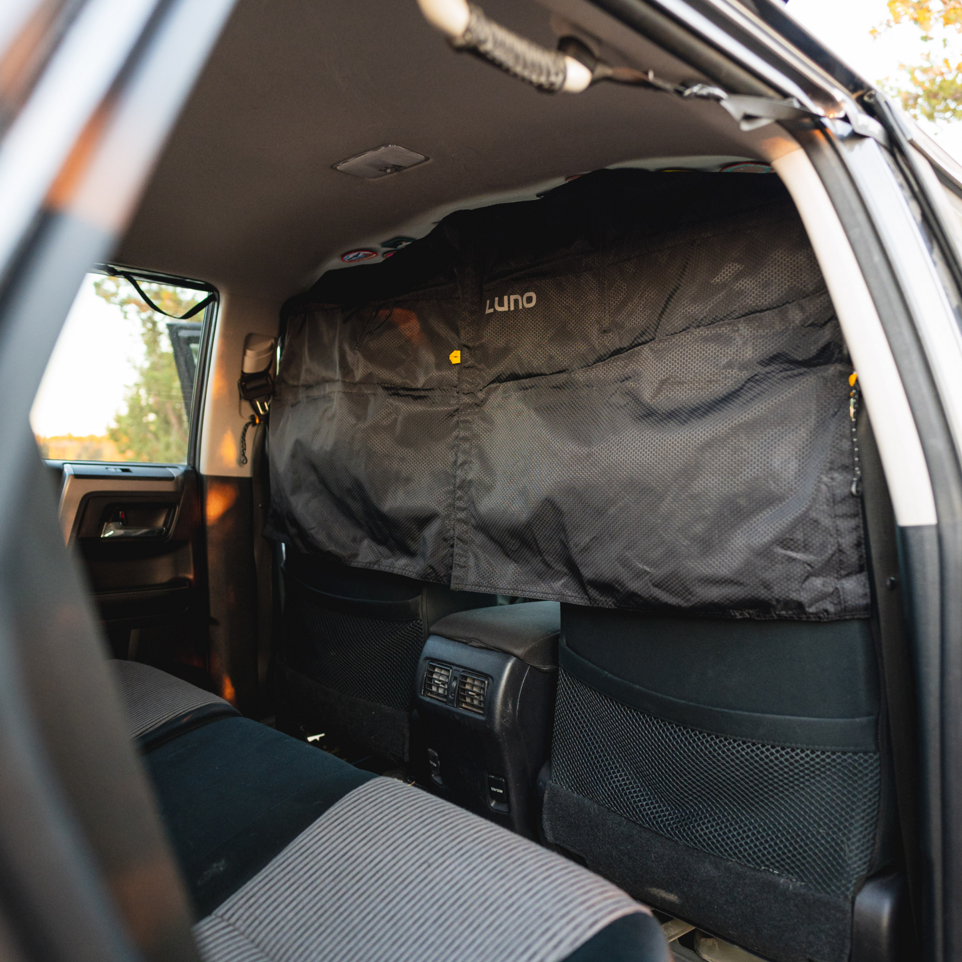 The image shows the backseat area of a car with a black Luno®-branded Car Privacy Curtain attached to the back of the front seats. The curtain has multiple compartments and covers the entire back of the seats, creating an ultimate sleeping space. The interior appears spacious and well-maintained.