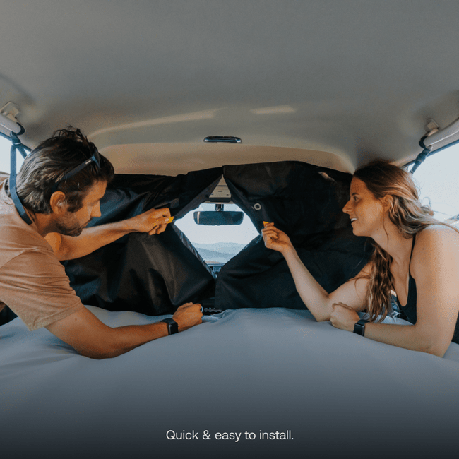 Two individuals are inside a vehicle, extending their arms to install pre-owned black Luno HQ Car Privacy Curtains on the rear windows. A wide view of the outside scenery can be seen through the windshield. The text at the bottom reads, "Quick & easy to install, available with USA shipping.