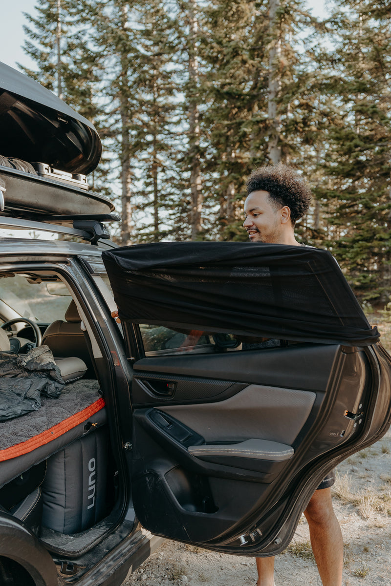 A person stands beside an open car door in a wooded area. The car is equipped with camping gear, including a sleeping bag and rolled-up mat visible through the door. Tall pine trees are in the background, giving a sense of outdoor adventure and nature.