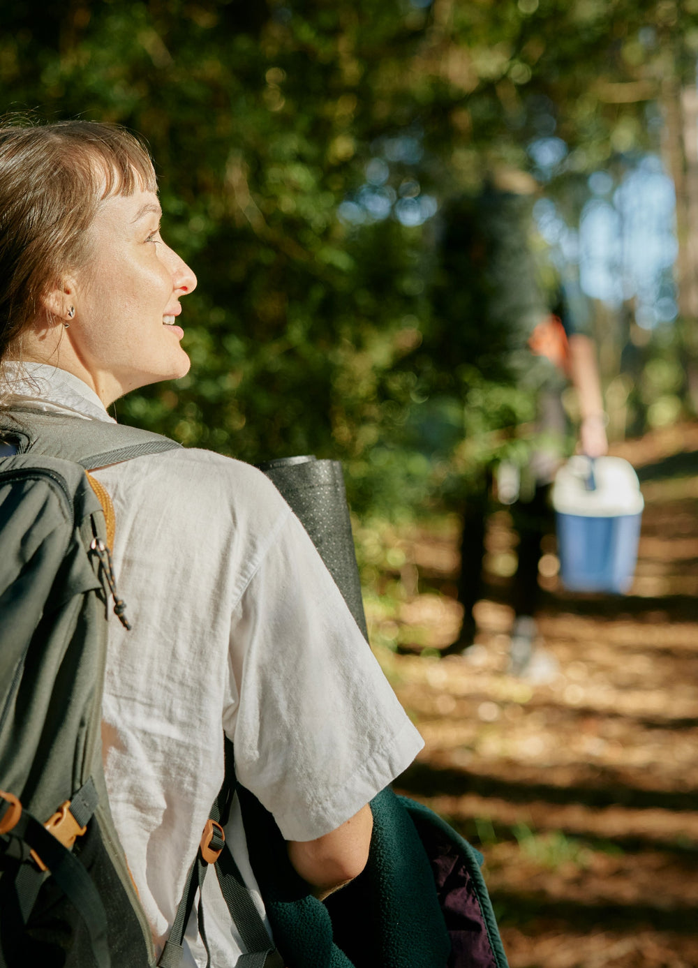 A woman with a backpack and rolled-up mat smiles while looking towards the trees in a sunlit forest. In the background, another person carrying a cooler walks along the path.