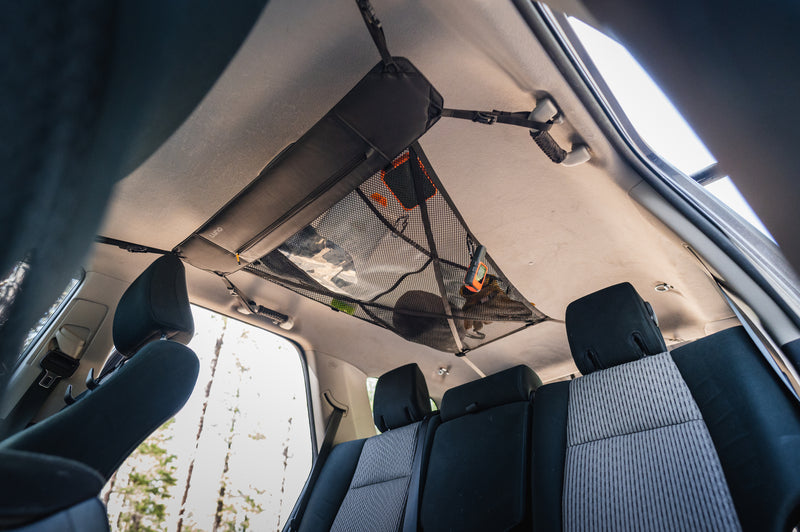 Interior of a car with a mesh storage net installed on the ceiling. The net contains some bags and small items. Black headrests and gray seats are visible, as well as partially open windows allowing daylight in. Trees can be seen through the windows.