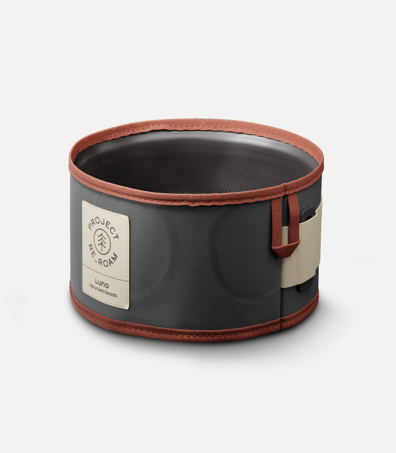 A round, collapsible dog bowl with a black exterior, a brown rim, and a beige strap with buckle. The bowl features a patch that reads "Project TREK ROAM" and "Luna" with a leaf logo on the front.