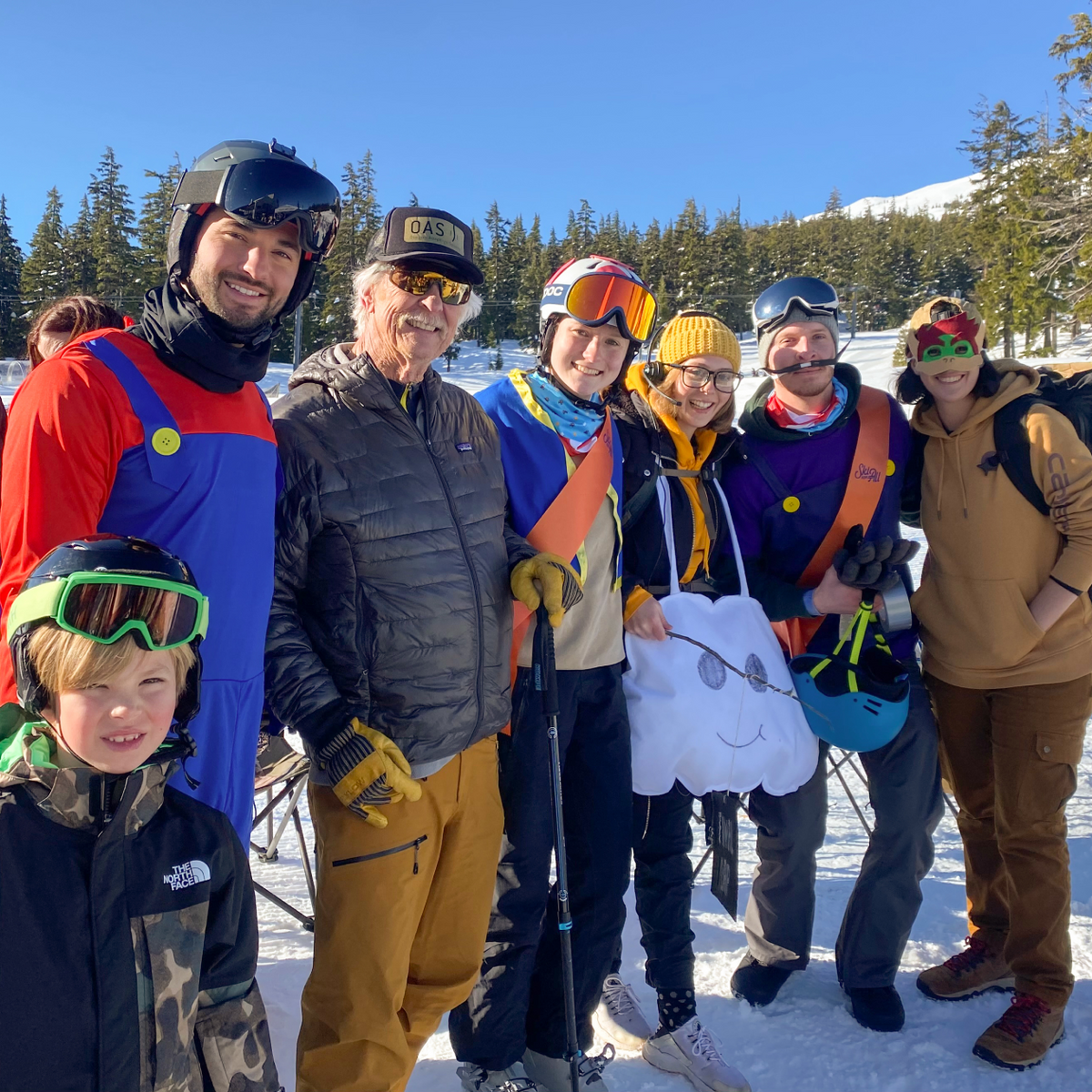 A group of seven people in winter clothing and ski gear pose for a photo on a snowy mountain. They are smiling and standing close together, with trees and clear blue skies in the background. Some are holding ski poles and wearing helmets or goggles.